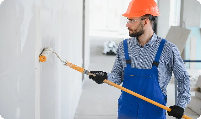 Man in blue overalls and orange hard hat painting a wall with a roller. He is focused, working in a bright, unfinished room.
