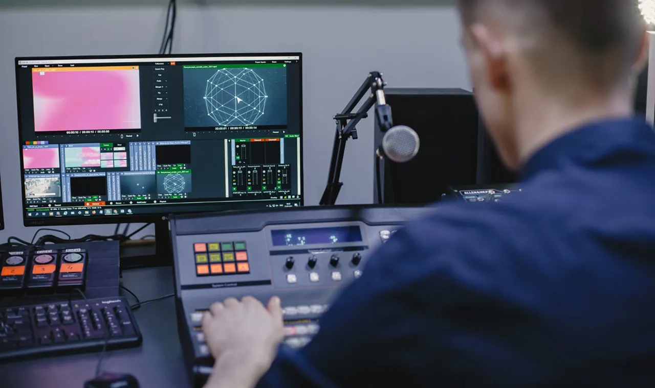 A person operates an audio mixing console in a studio, with multiple monitors displaying graphics and controls. The scene conveys focus and professionalism.