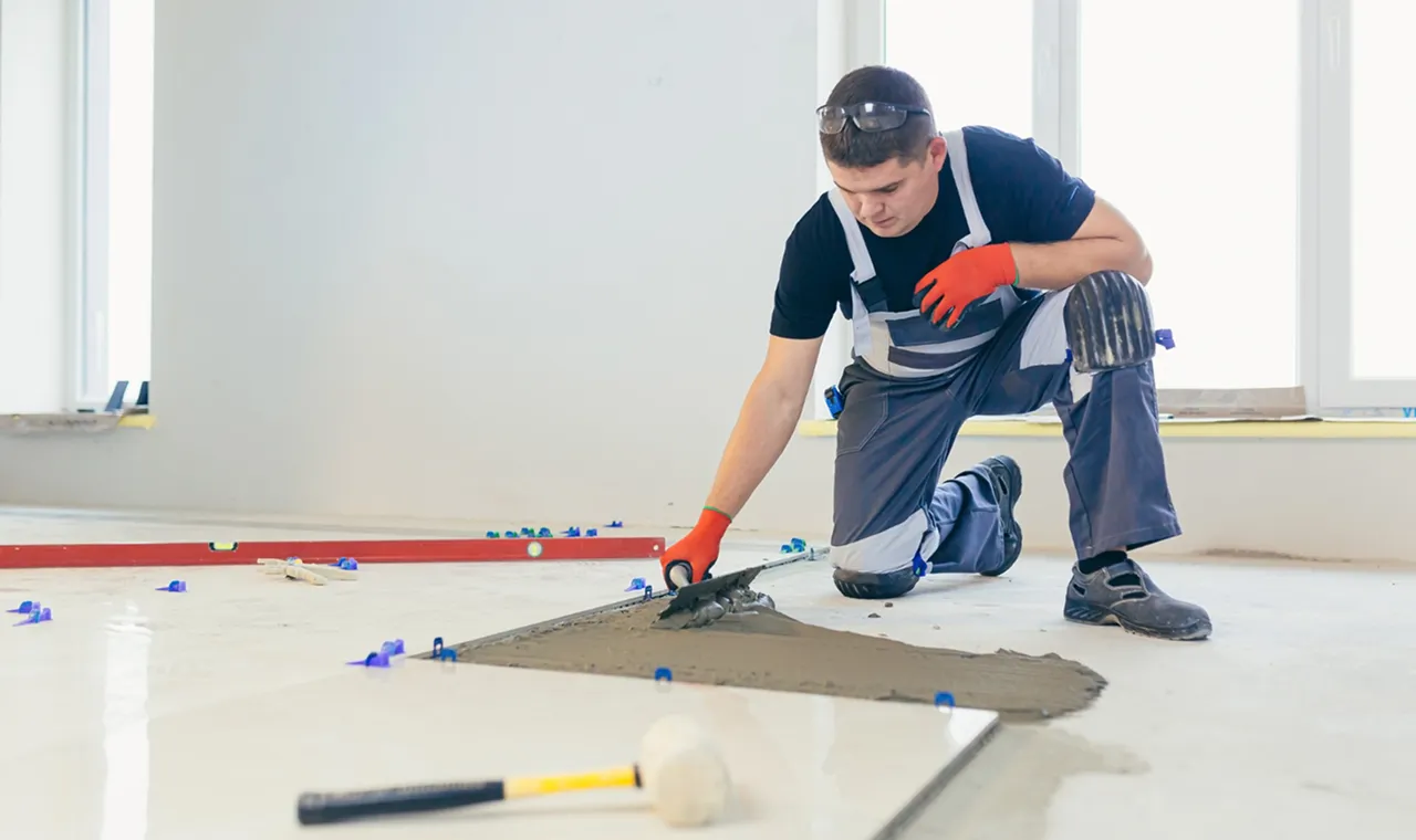 A construction worker in gloves and kneepads is kneeling, smoothing concrete on a floor with a trowel. The scene conveys focus and precision.