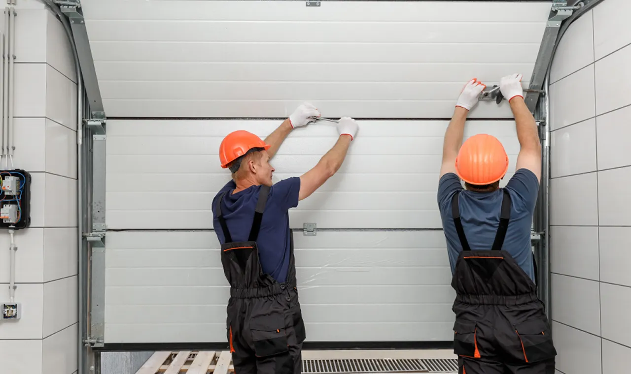 Two workers in orange hard hats and gloves install a garage door, focused on ensuring it fits correctly within the frame.