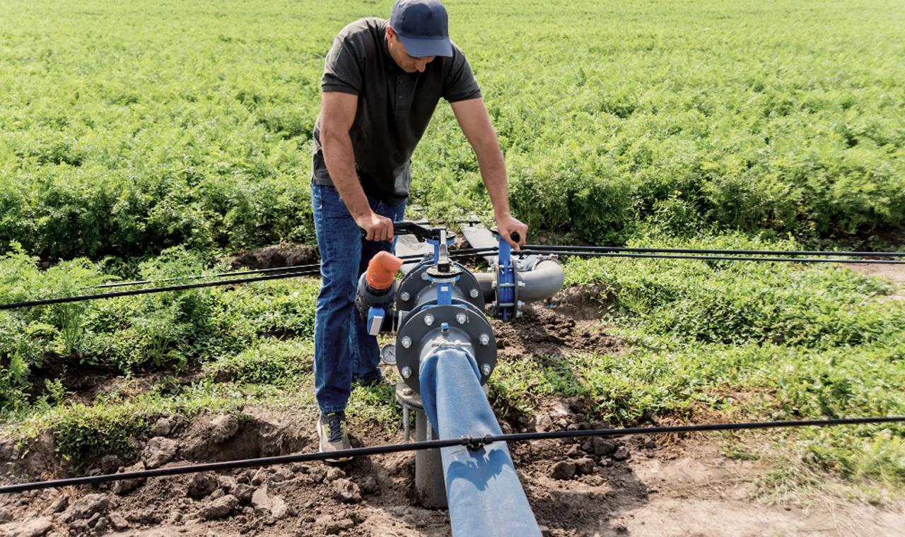 Man in blue cap and jeans adjusts irrigation pipe in green field, focusing on control valve. Bright, sunny day highlights agricultural work scene.