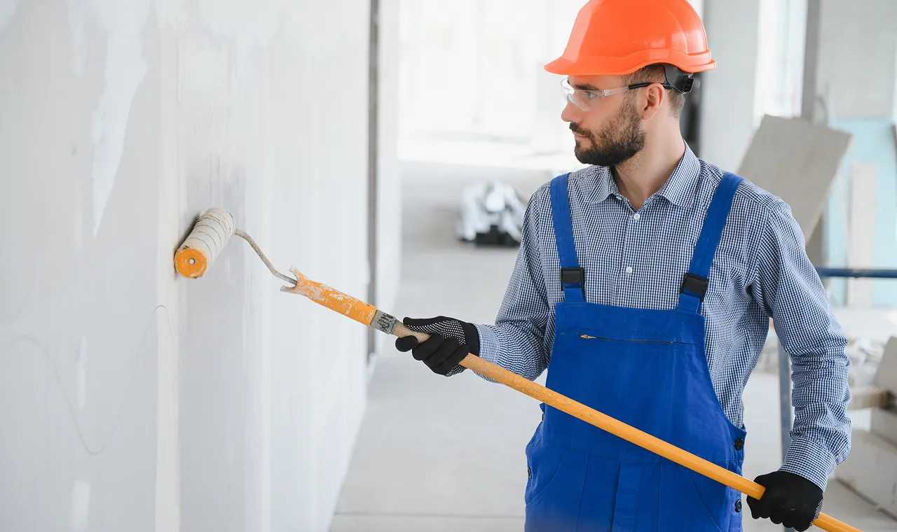 Man in blue overalls and orange hard hat painting a wall with a roller in a bright, unfinished room.